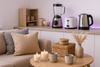 Realistic photo of a cozy, modern home interior featuring both home and kitchen essentials — soft cushions, scented candles, storage baskets, ceramic mugs, and neatly arranged decor. The kitchen area includes smart appliances like a sleek mixer grinder, toaster, and electric kettle. The background has a clean white and light purple neon glow theme, giving a fresh, elegant, and organized look. The scene feels warm, inviting, and stylish — highlighting comfort, innovation, and a modern lifestyle vibe